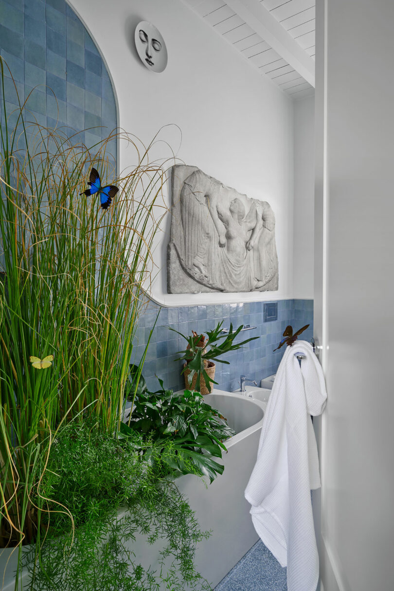 A bathroom with blue tiled walls, lush green plants around the bathtub, a butterfly, and decorative wall art.