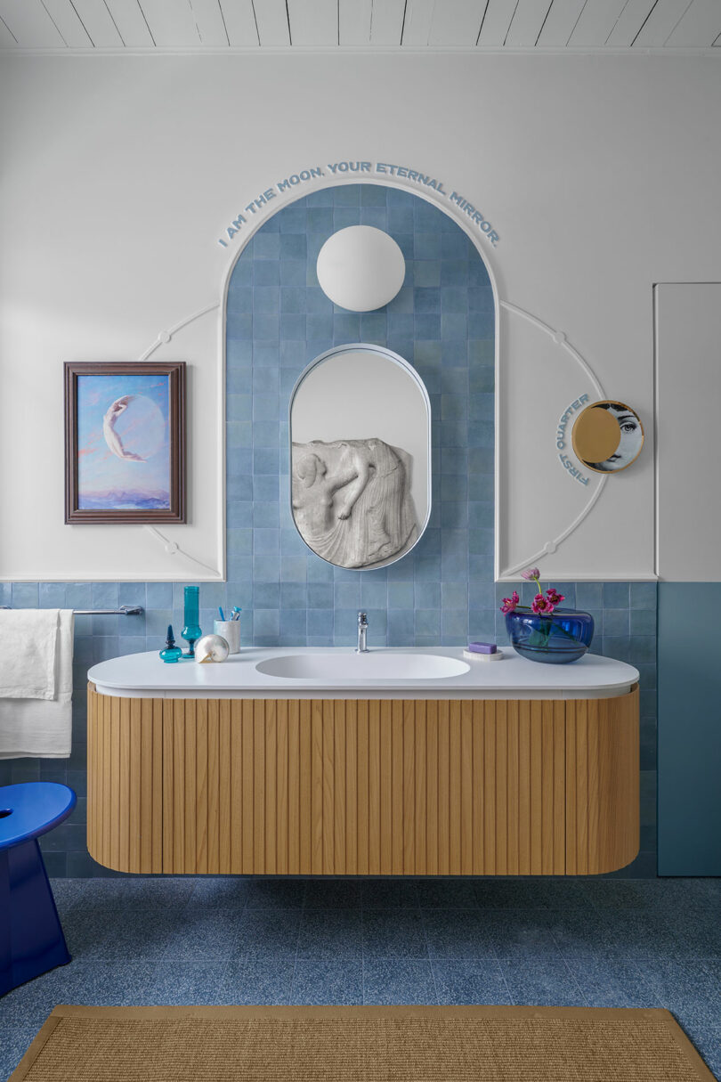 Modern bathroom with a curved wooden vanity, blue tiles, and a round mirror. Wall inscription reads, 