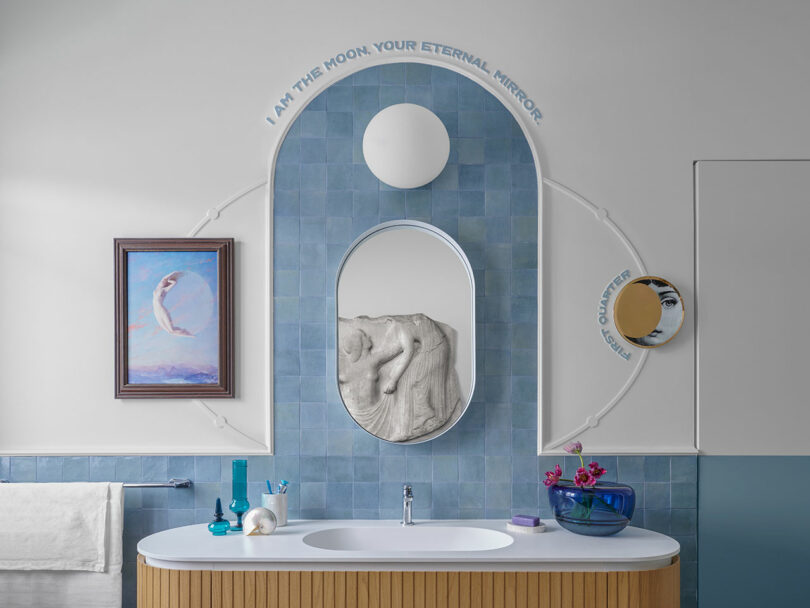 Bathroom with blue tiles, an oval mirror, a circular light, artwork, and text on the wall. A wooden vanity with white countertop holds toiletries and flowers.