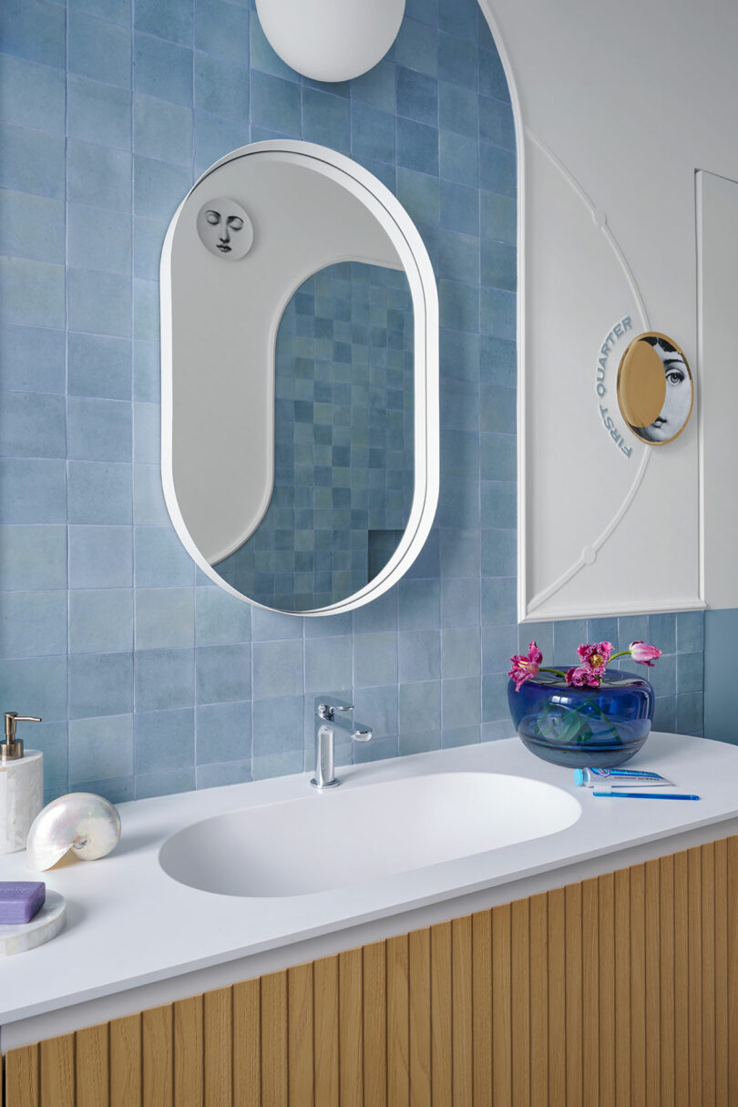 Bathroom with wood vanity, oval mirror, blue tiles, and a white countertop. A small bowl with pink flowers is on the counter next to soap and a seashell.