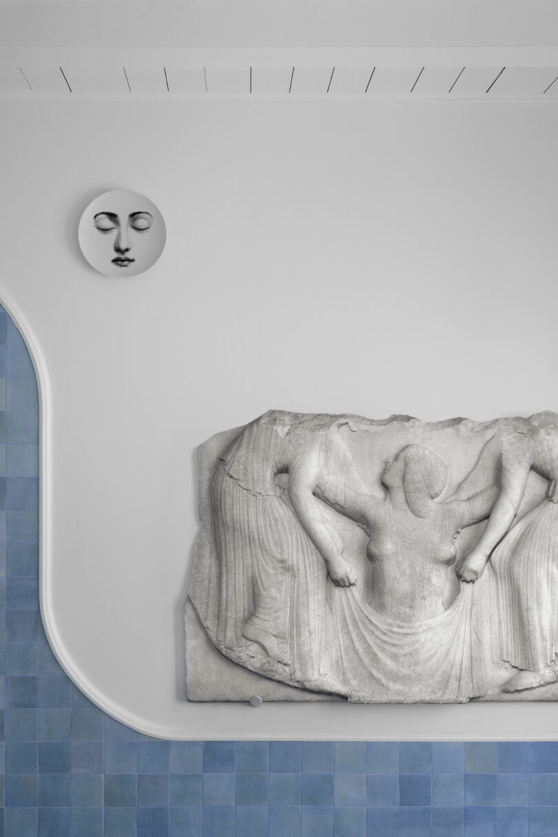 A stone relief sculpture depicting human figures is mounted on a white wall. Above it is a round wall ornament with a face, and below is a blue-tiled surface.