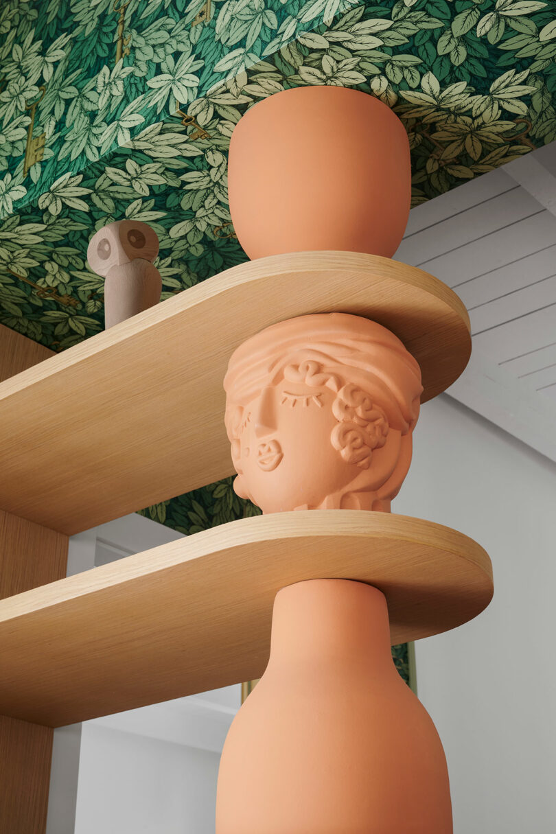 Terracotta vases with carved faces are placed on wooden shelves against a leafy ceiling design.