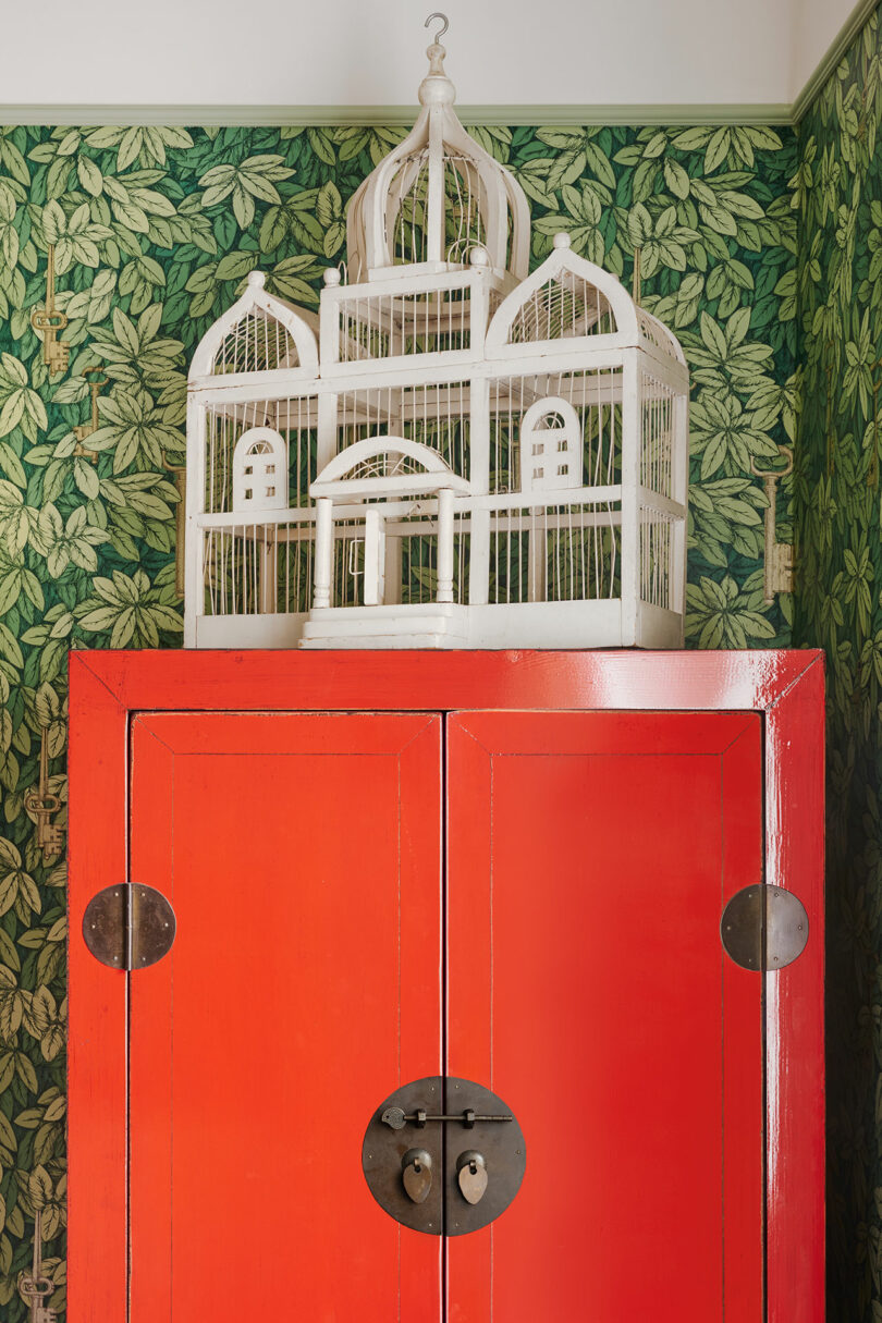 A red cabinet with a round lock stands against green leaf-patterned wallpaper. A decorative white birdcage sits on top.