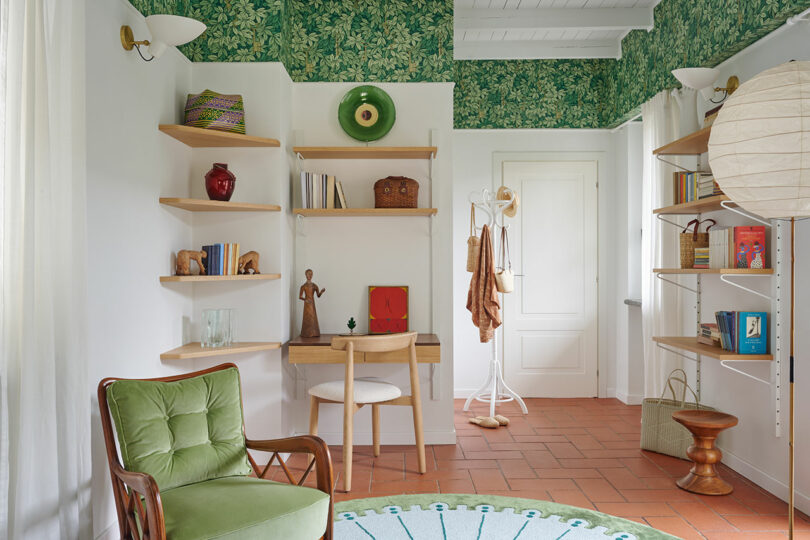 A cozy room with green foliage wallpaper, a small desk with a chair, open shelves with books and decor, a wooden floor, and a green cushioned chair in the foreground.