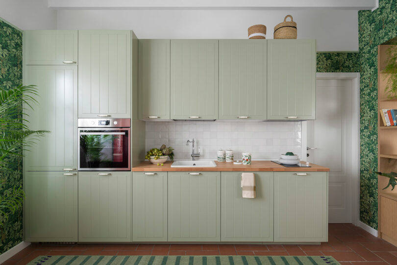 A kitchen with green cabinets, a wooden countertop, built-in oven, and leafy wallpaper. A striped rug and various kitchen items are visible on the countertop.