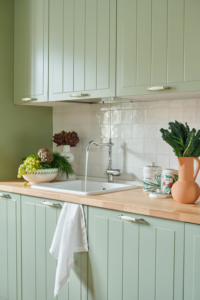 A kitchen with light green cabinets, a white sink, and a wooden countertop. Fresh produce is on the counter, including grapes, pineapple, and kale. A white towel hangs by the sink.