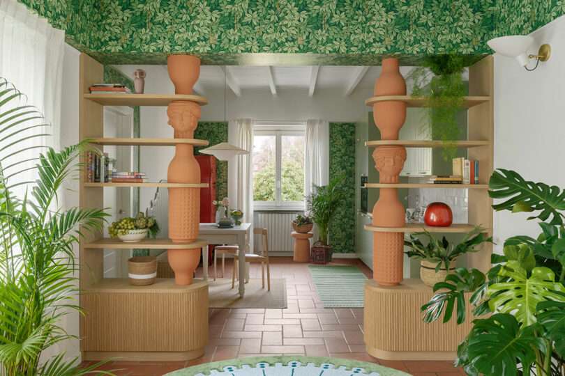 Room with green leaf-patterned wallpaper, terracotta flooring, large planters, and wooden shelves separating dining and living areas. Bright window view and various plants enhance the space.