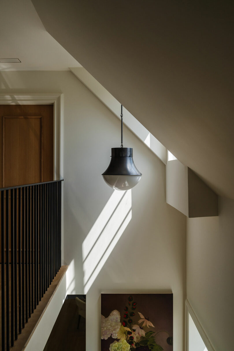 A stairwell with a hanging black pendant light, slanted ceiling, and wooden door features a floral painting. Sunlight creates diagonal shadows on the white walls.