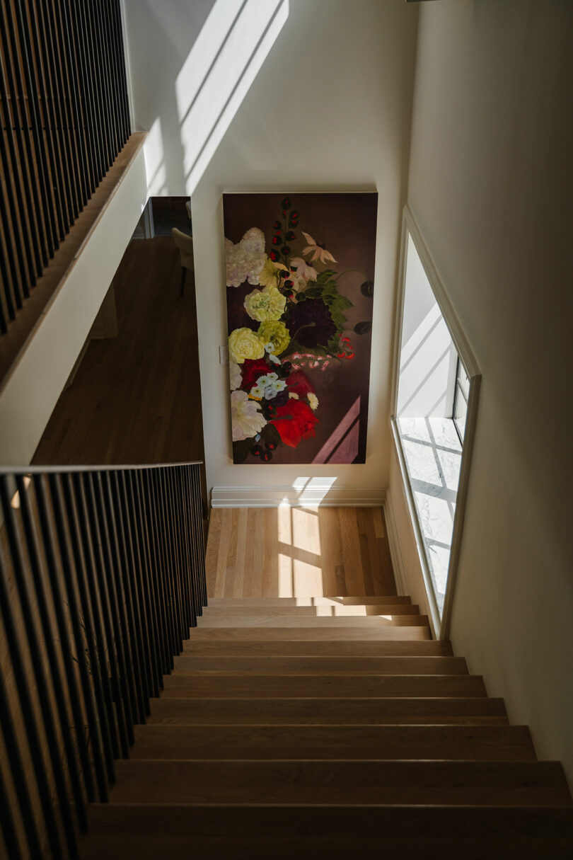 Staircase with wooden steps and metal railing, adjacent to a large floral painting of roses. Natural light from a window casts delicate shadows on the wall and floor.