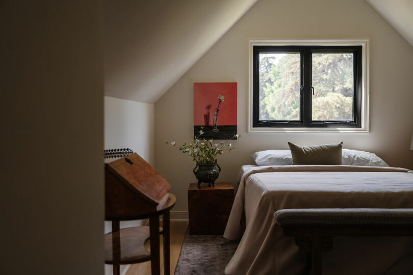 Cozy attic bedroom with a sloped ceiling, featuring a bed, a painting by Louis on the wall, a window with a view of trees, and charming touches like a small table and wooden desk.