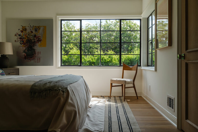 A sunlit bedroom with a large window showcases a bed, chair, and artwork on the wall. A striped rug covers the wooden floor, weaving together the serene ambiance.