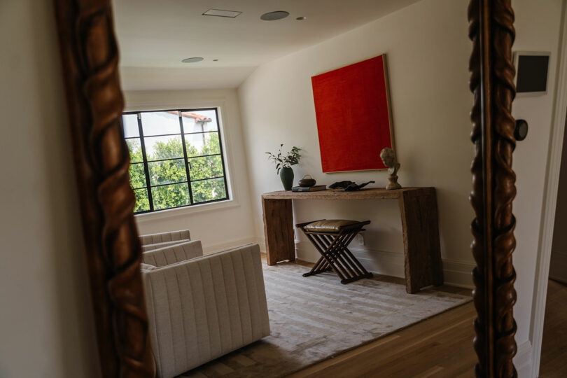 A cozy room with beige sofas and a wooden table, where a red painting by Louis adds a touch of elegance. A potted rose plant sits on the table near a window, offering a view of lush greenery.