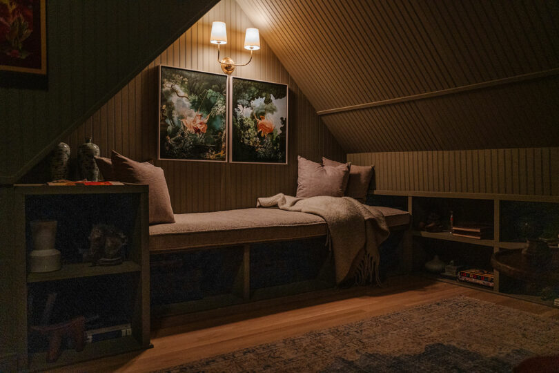 A cozy attic reading nook features a cushioned bench with pillows and a blanket, surrounded by bookshelves. Two nature-themed paintings and a wall lamp decorate the slanted wooden wall.