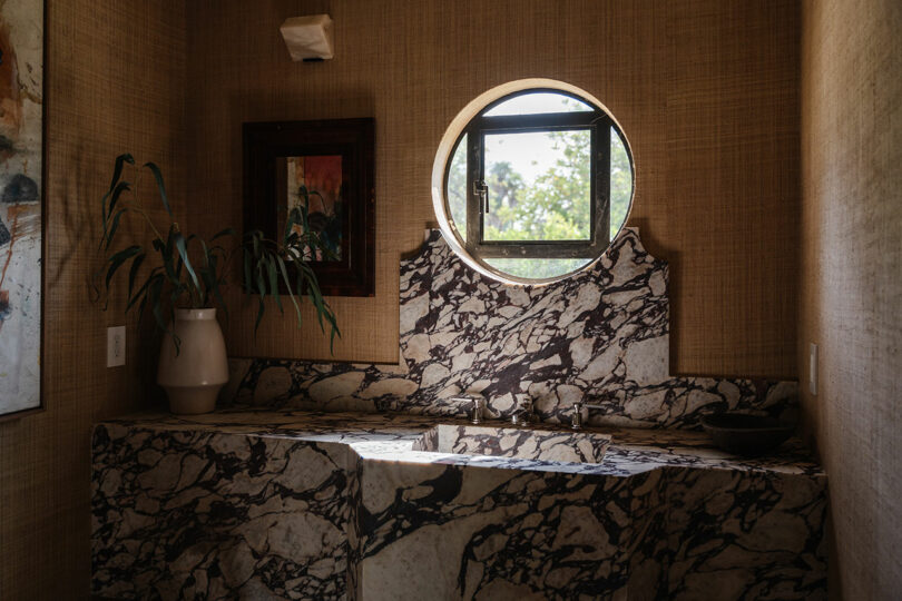 Bathroom with a marble sink, a round window, and a potted plant. Framed artwork complements the textured walls, adding an elegant touch.