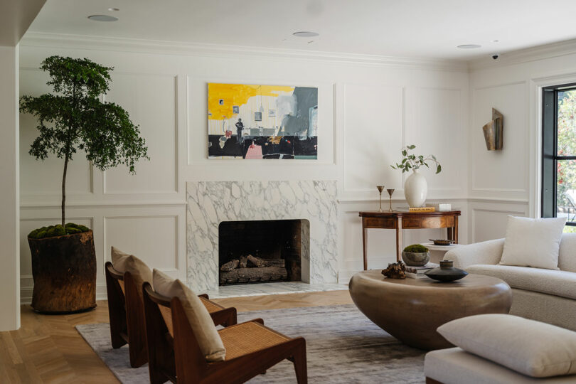 A modern living room with a marble fireplace, abstract art above, wooden furniture, a large potted plant, and a round coffee table on a rug creates an inviting atmosphere.