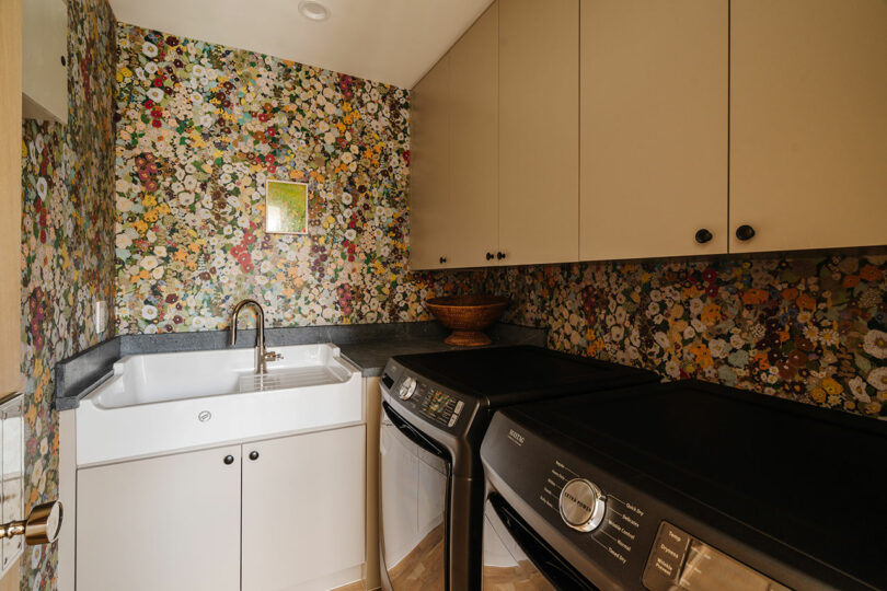 The laundry room, a charming space features a sink, white cabinets, and a sleek black washer and dryer. The walls are adorned with floral wallpaper, and a rustic wooden bowl rests elegantly on the counter.