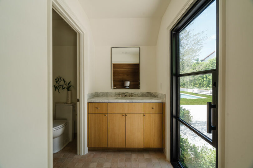 A small bathroom with a wooden vanity and marble countertop. A mirror is above the sink, and a window on the right offers a view of greenery outside. To the left, a toilet is partially visible next to a plant.