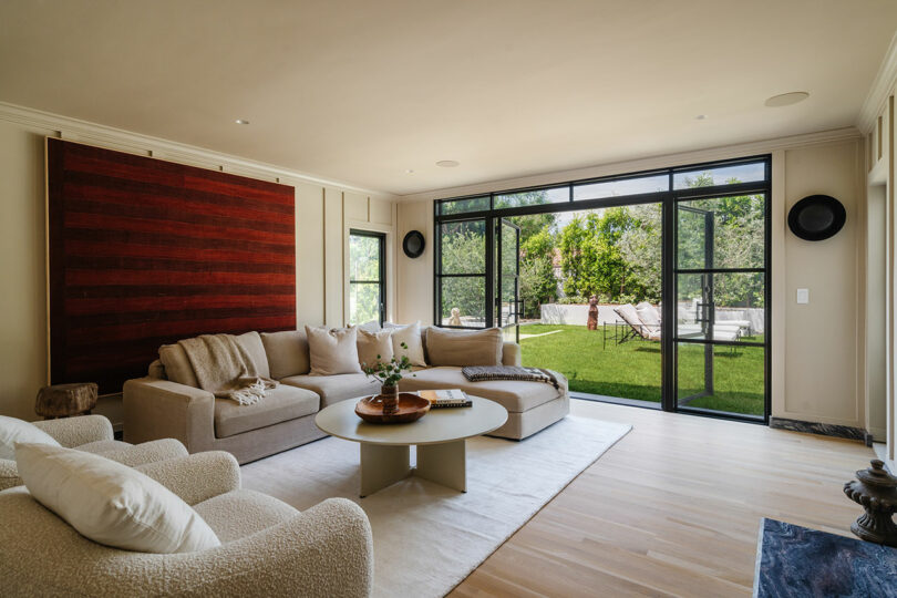 A modern living room features a beige sectional sofa and a white armchair, with large glass doors opening to a lush green lawn.