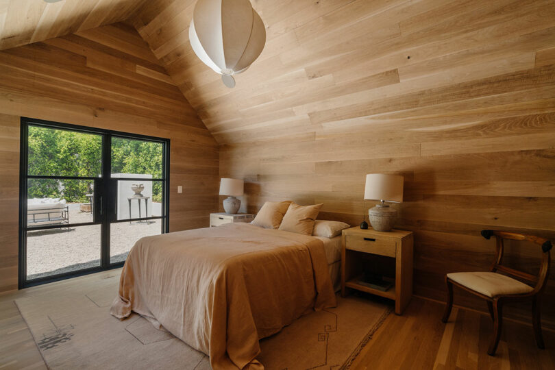 The bedroom, with its wooden walls and ceiling, features a bed draped in beige linens. Flanking each side are two bedside tables with lamps. A chair sits by the large window offering serene views of the outdoors.