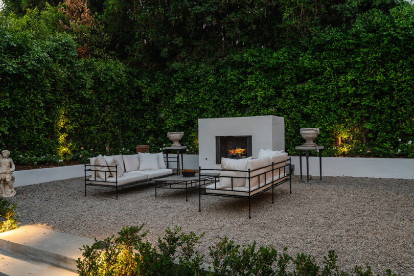 The outdoor seating area features two white couches and a cozy fireplace, surrounded by lush greenery and gravel ground.