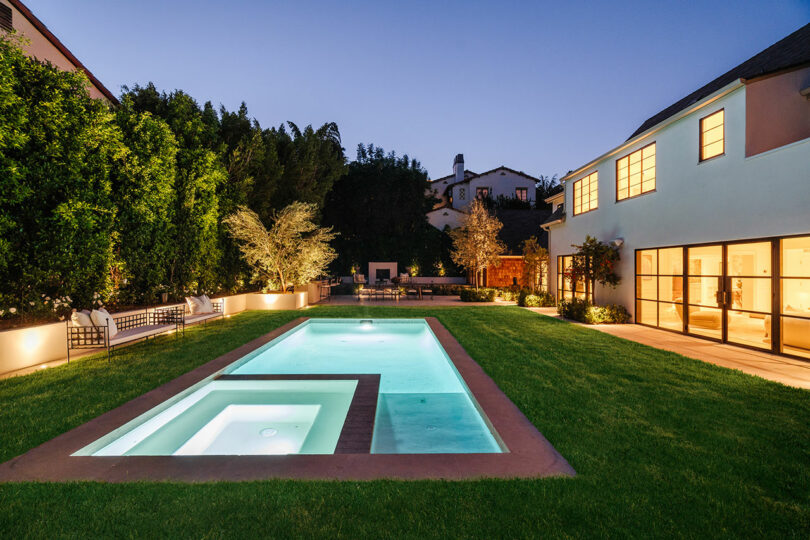 At dusk, this modern house boasts a well-lit backyard with a rectangular pool and hot tub, nestled amid grass and trees.