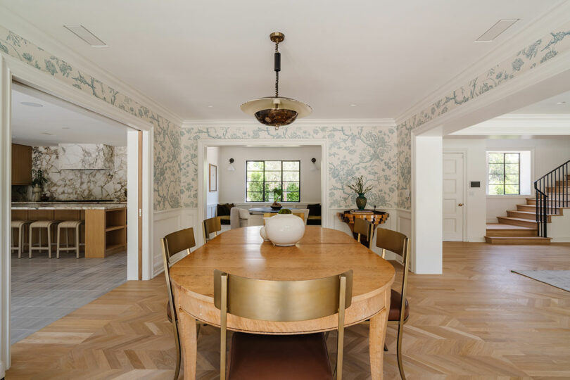 The dining room, adorned with wooden table and chairs, features floral wallpaper and offers a charming view into the kitchen and living area. Stairs are visible to the right, adding a touch of elegance reminiscent of classic design.