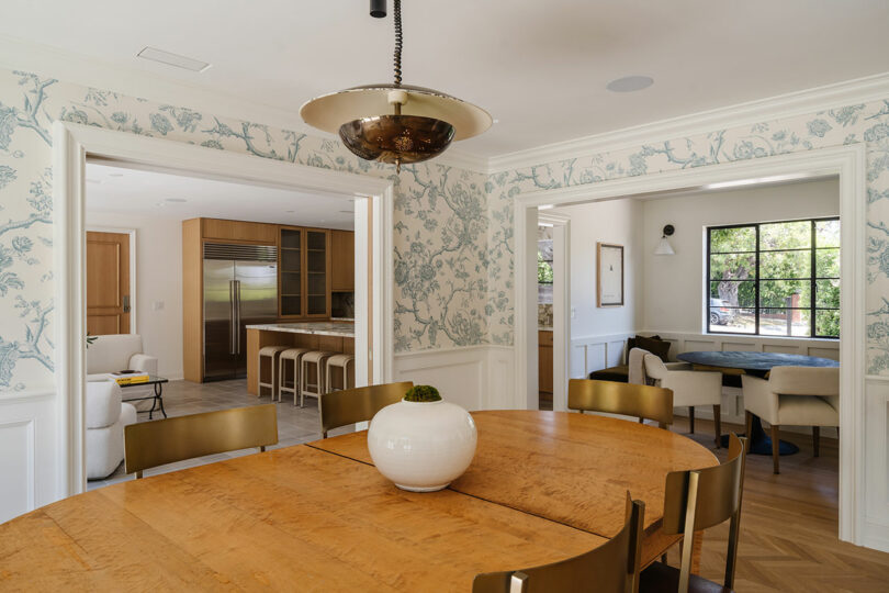 A dining room with a wooden table and elegant gold chairs, adjacent to a kitchen and living area. The space exudes charm, with floral wallpaper and a large window bathing everything in rosy natural light.