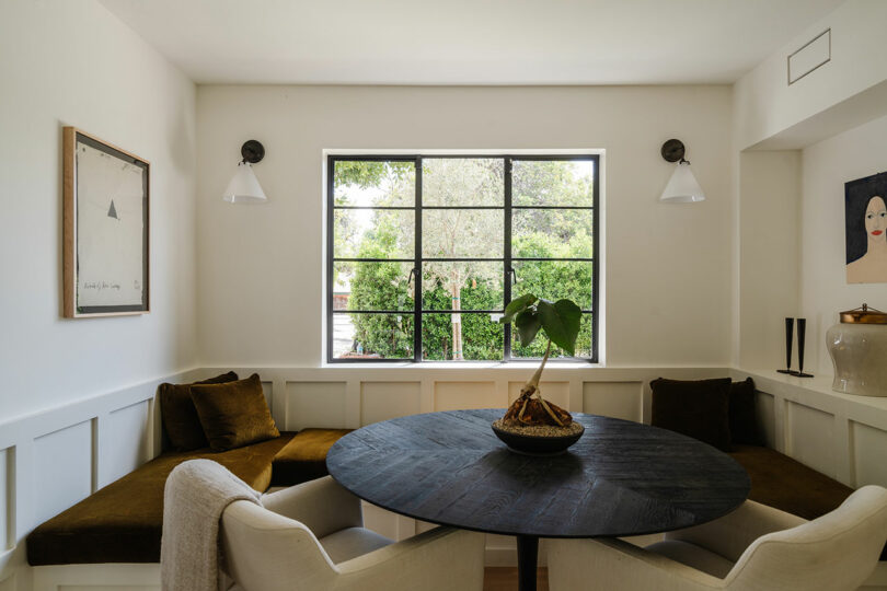 Minimalist dining area with a round black table, white chairs, and a rose centerpiece. Two wall artworks and a large window with a view of greenery complete the space, adding an elegant touch.