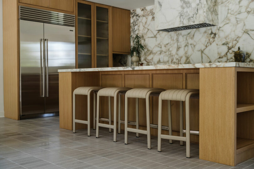 Modern kitchen with a marble backsplash and stainless steel fridge. Wooden cabinets add warmth, while four stools line the island with a countertop matching the elegant backsplash.