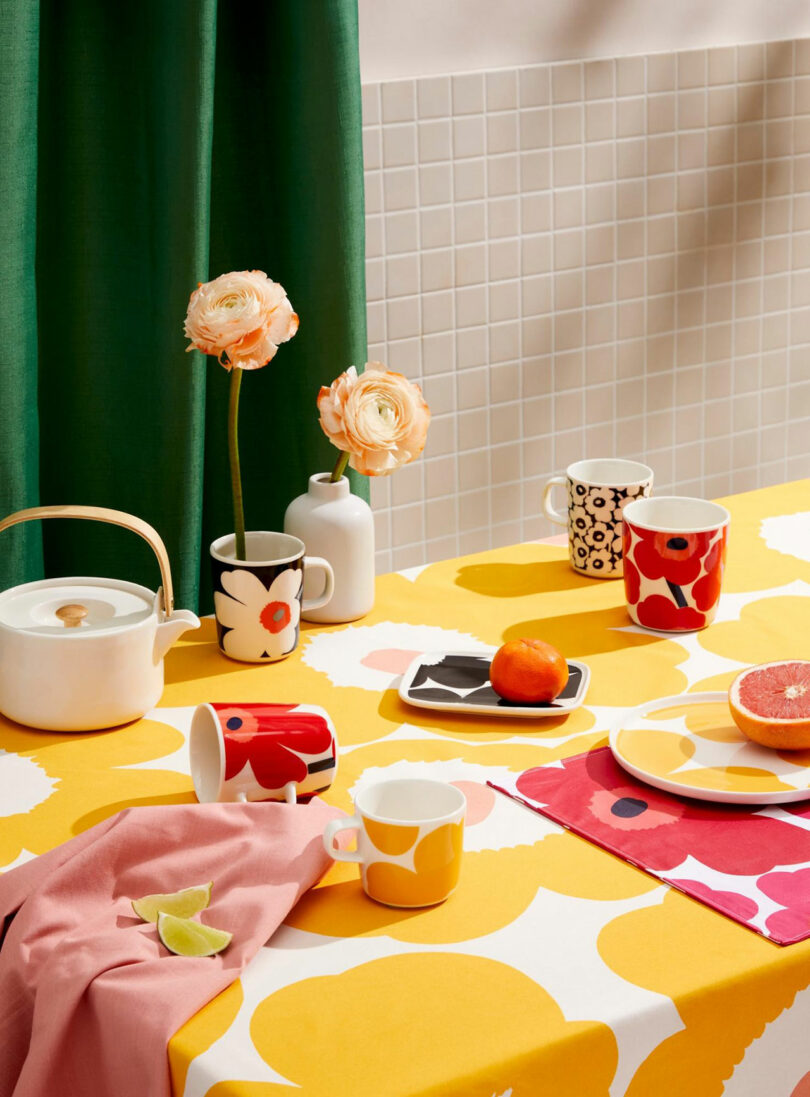 A colorful kitchen table adorned with mugs, a teapot, a vase of flowers, and juicy fruit evokes the vibrant artistry of Giulio Barresi. Bright floral patterns dance on the tablecloth and cups, set against a tiled wall and lively green curtain backdrop.