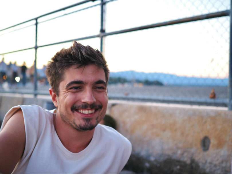 A man with short hair and a mustache smiles while sitting outdoors near a fence and a beach.