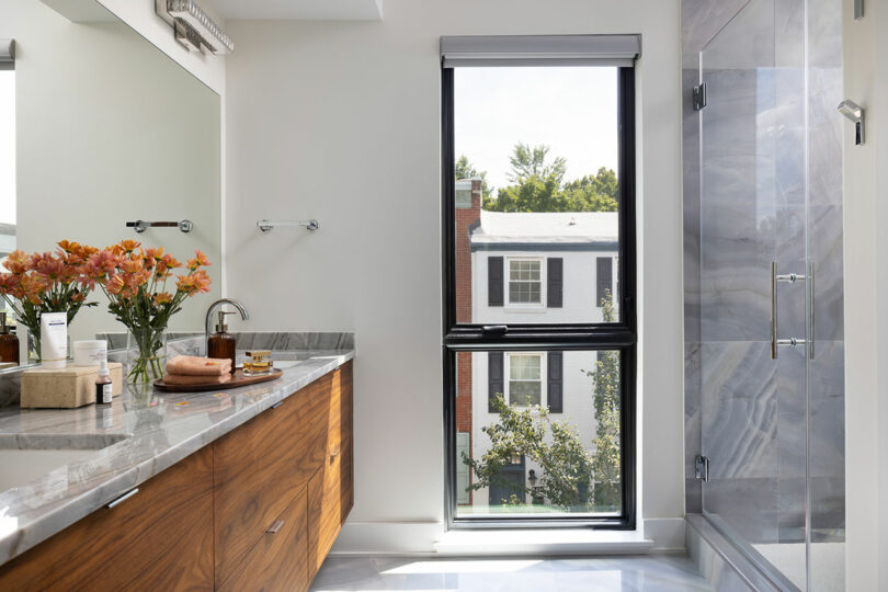 Modern bathroom featuring a wooden vanity, marble countertop, and a vase of flowers. A large window offers views of the city skyline, while a glass-enclosed shower stands elegantly to the right.