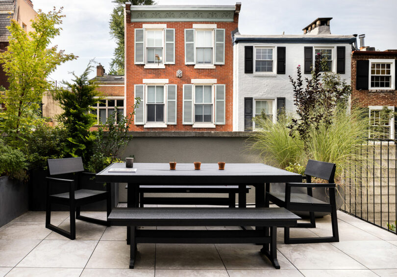 View of a modern patio with a black dining set, lush plants, and a backdrop of charming brick and white townhouses.