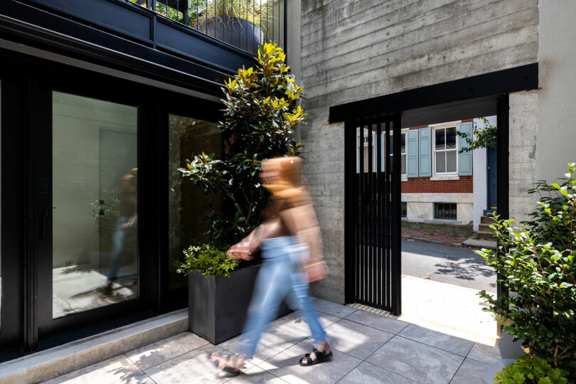 A person strolls beside a window adorned with plants, nestled in a courtyard where gnome architecture meets the robust charm of concrete and brick.