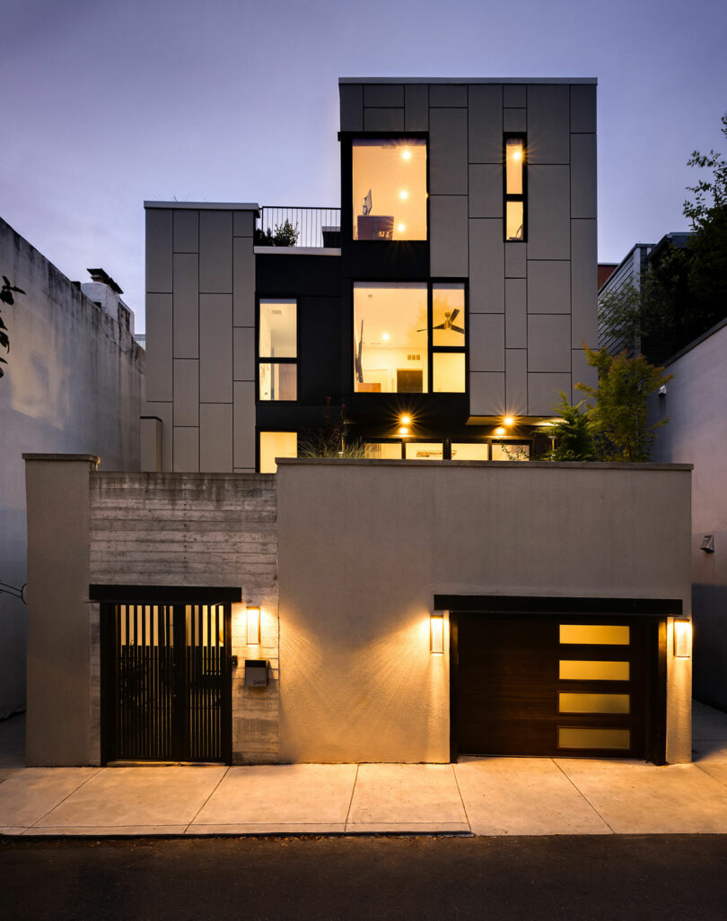 This modern multi-story building features a flat roof, large windows, and a garage. The exterior blends concrete and paneling, beautifully illuminated by several lights at dusk.