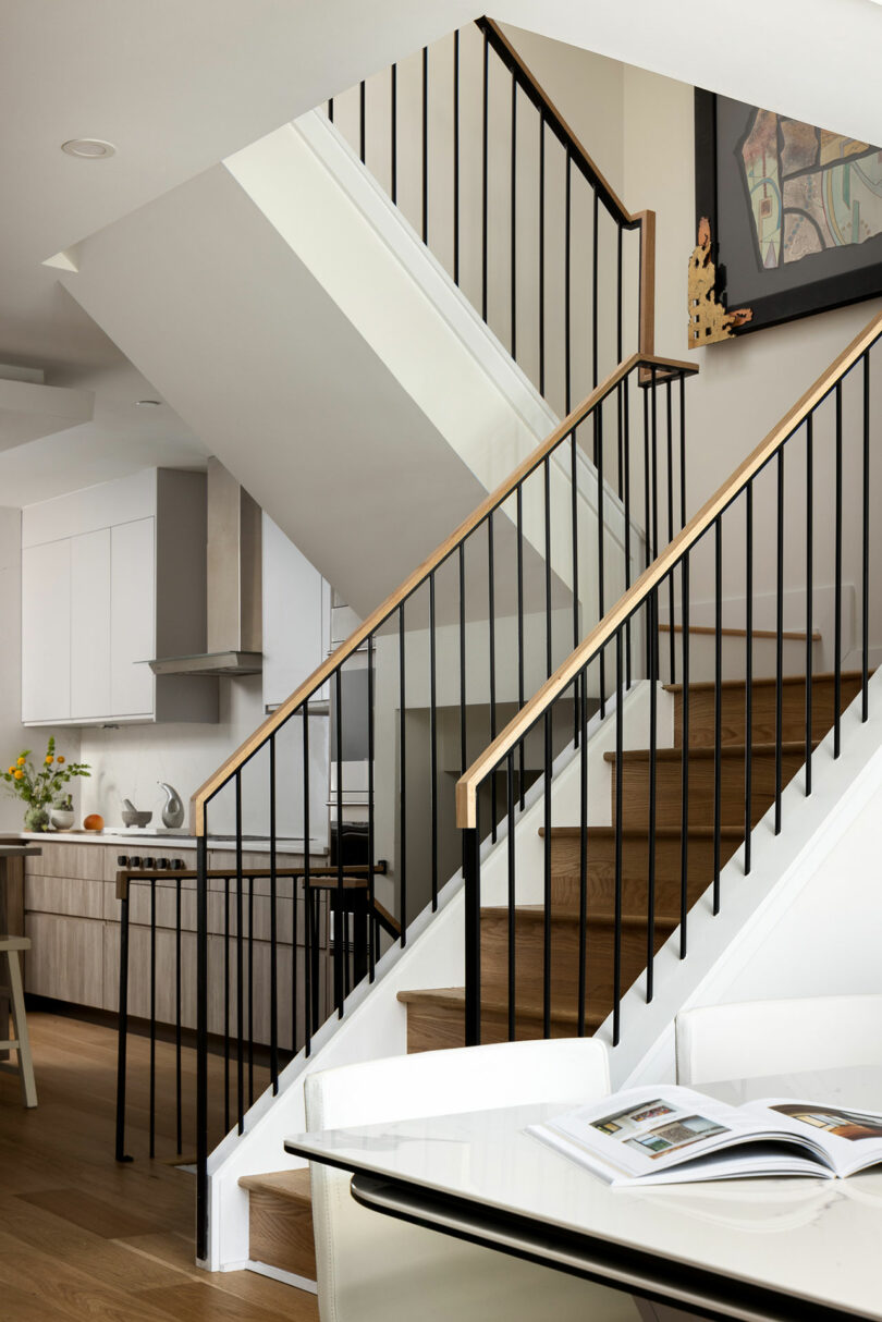 The modern staircase features sleek black railings and elegant wooden steps, perfectly complementing the light-filled home interior. In the background, the kitchen area beckons, while an open book graces the white table in the foreground.