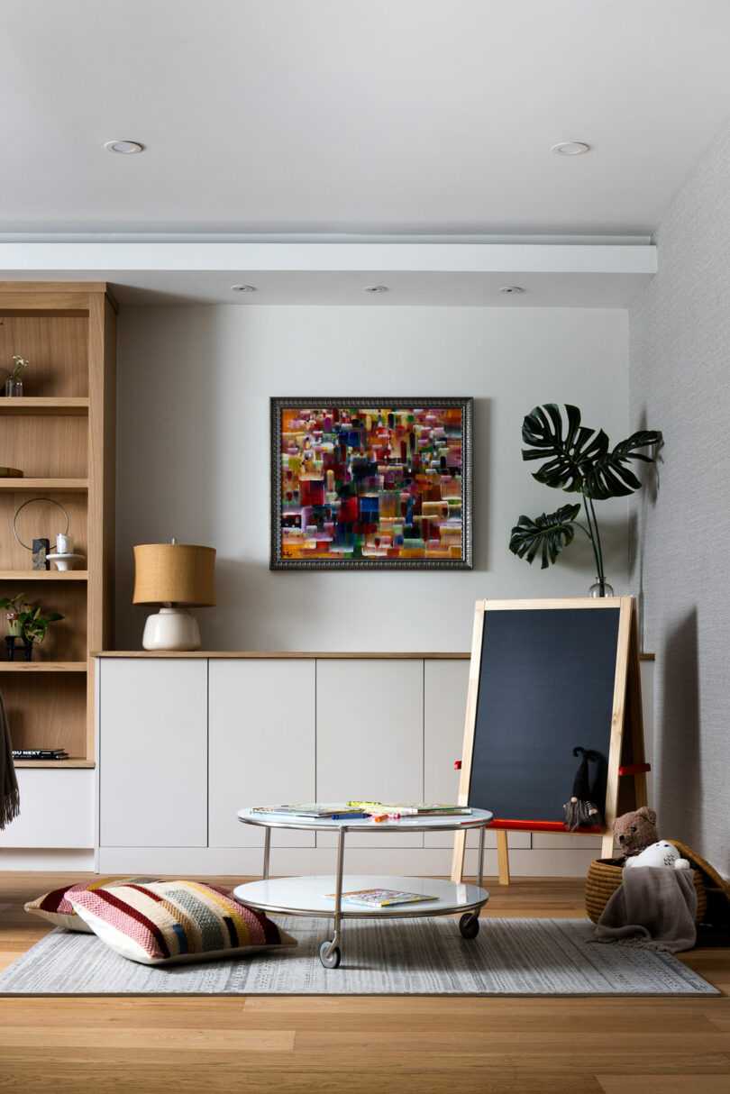 This modern playroom features a chalkboard easel, colorful cushions, and a low table with magazines. A framed abstract painting adorns the wall, while shelves hold a lamp and charming decorative items.