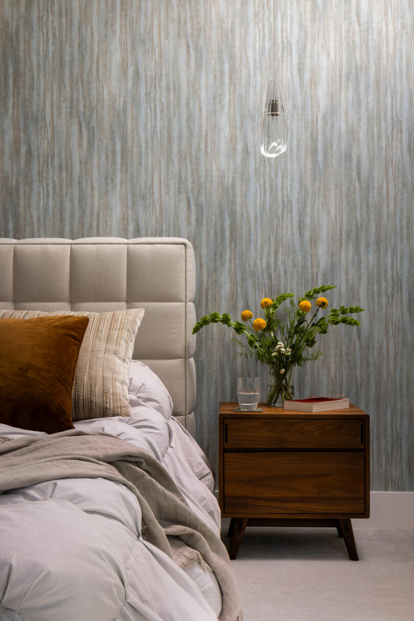 The bedroom scene features a charming tufted headboard, a side table adorned with a vase of yellow flowers, and a glass of water beside stacked books. A single hanging light fixture adds warmth above.