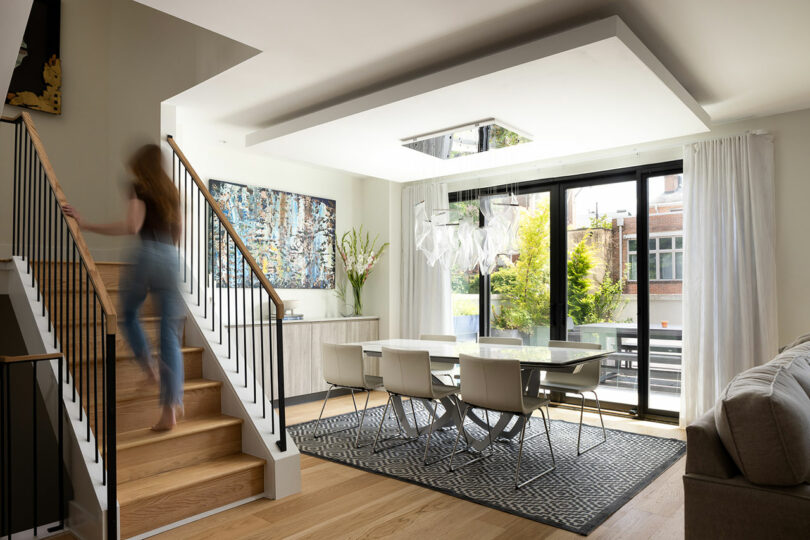 A woman ascends the stairs in a modern living space, designed by Gnome Architects, featuring a dining area with a glass table, white chairs, large windows, and captivating wall art.