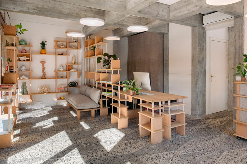 A modern room with open shelves, a desk, a couch, and decorative plants. The ceiling features circular lights, and the floor is made of textured tiles