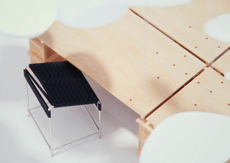 A black woven chair with a metal frame is placed next to a wooden table made of panels on a light floor. White abstract shapes hang in the foreground