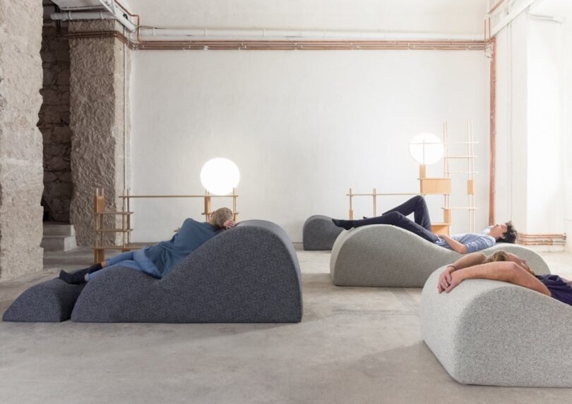 People lying on uniquely shaped cushioned furniture in a minimalist room with exposed pipes and soft lighting