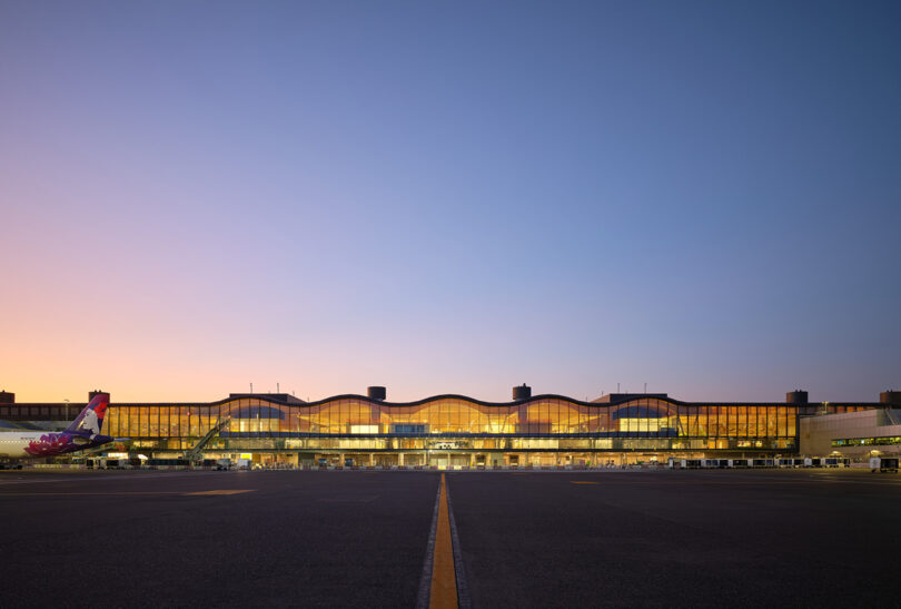 As dusk descends, the exterior view of the ZGF-designed airport terminal comes alive, with a plane parked on the left and the building beautifully illuminated under a clear sky.