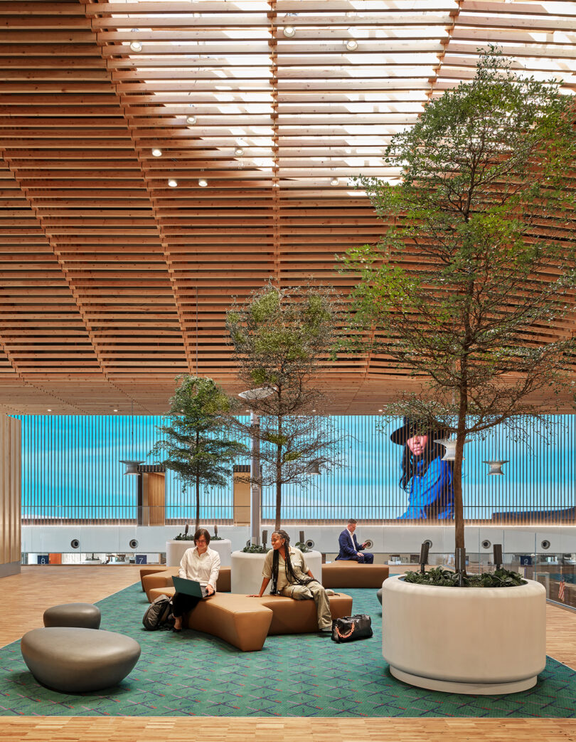 The modern interior, designed by ZGF, features a wooden ceiling and trees in planters. People relax on cushioned seats as a large digital screen showcases an image of a person in blue.