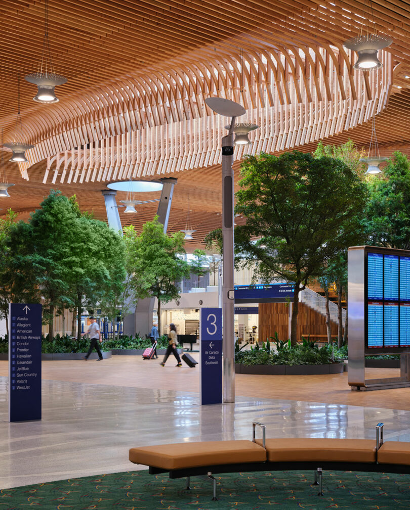 A modern airport terminal designed by ZGF features a stunning wooden ceiling, lush indoor trees, ample seating, and a sleek digital departure board. Passengers leisurely stroll through the airy space with luggage in tow.