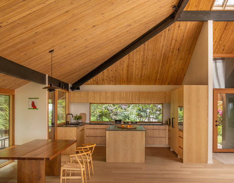 A modern kitchen with wooden cabinetry and ceiling, a central island, and large window overlooking greenery. A wooden dining table with chairs is in the foreground.