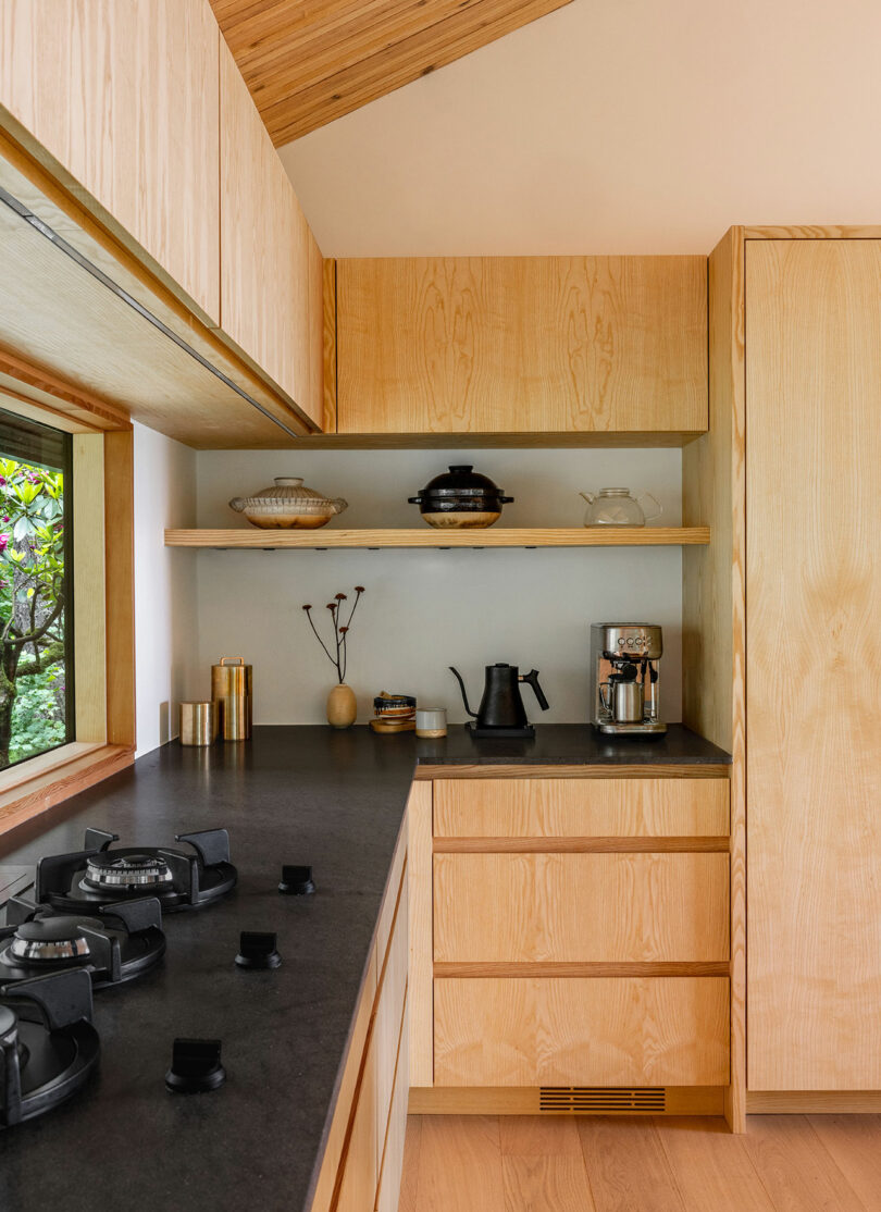 Modern kitchen with light wood cabinets, black countertop, gas stove, coffee maker, kettle, and decorative items on open shelf. Large window with greenery outside.