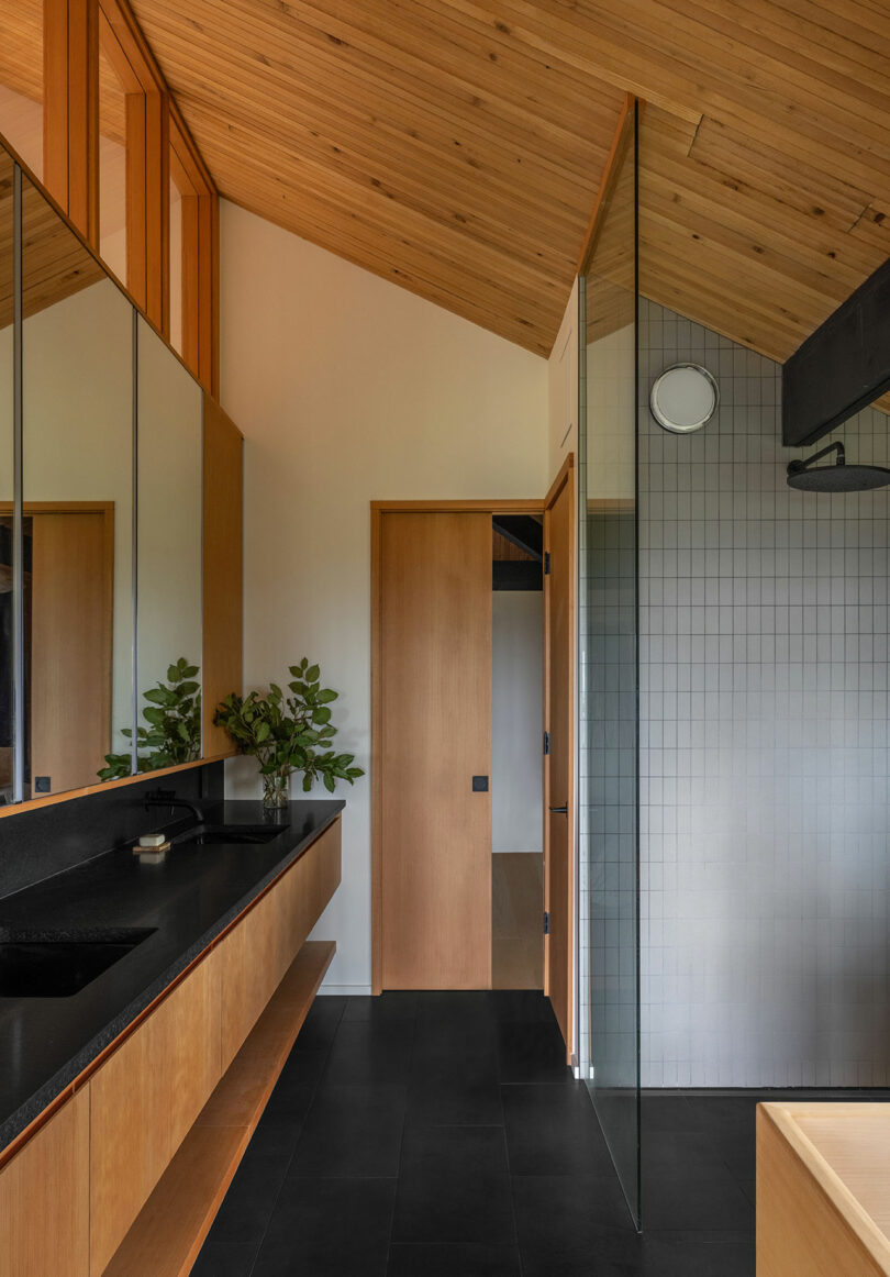 Modern bathroom with wooden accents, large mirror, black countertop, glass shower partition, and black floor tiles.