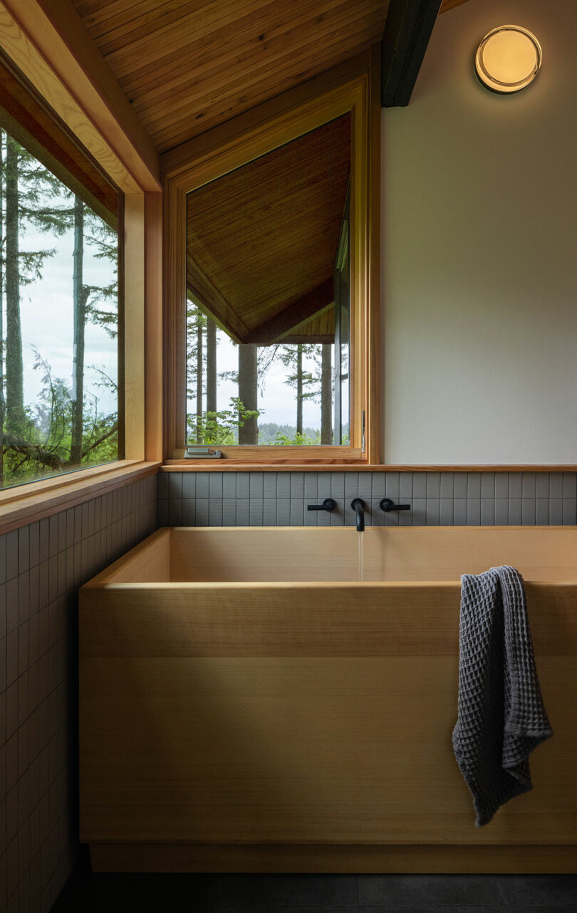 A wooden bathtub with a gray towel draped over the edge is positioned by large windows, offering a view of trees. The room has tiled walls and a ceiling light.