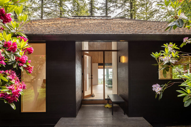 A dark wooden house with large windows and a central glass door, surrounded by blooming pink flowers and tall trees.